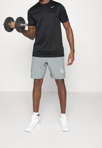 Černé tričko Nike s krátkým rukávem, spárováno se světle šedými šortkami se bílým logem. Drží černou činku a má na sobě bílé sportovní boty.