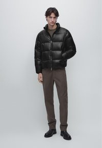 Młody mężczyzna w czarnej pikowanej kurtce, brązowych spodniach i czarnych botkach do kostek, stojący na tle jednolitego jasnoszarego tła.