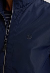 Navyblauwe jasje met rits, gemaakt van soepel, lichtgewicht materiaal met een subtiele glans. Beschikt over een logo op de borst en een opstaande kraag.