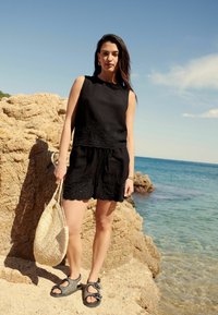 Svart ärmlös topp med broderade detaljer, parat med svarta shorts, håller en vävd väska, står på en stenig strand vid vattnet.