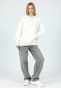Sudadera blanca con capucha, hecha de algodón, con un bolsillo frontal y puños acanalados, combinada con vaqueros de mezclilla gris claro de corte holgado y zapatillas blancas.