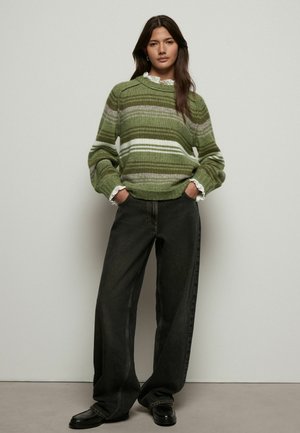 Maglione a righe verdi con sfumature variabili e accenti bianchi, abbinato a jeans larghi scuri e scarpe nere. Materiale a maglia testurizzato.