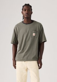 Ριγέ κοντομάνικη μπλούζα σε σκουροπράσινο και μπεζ, με στρογγυλή λαιμόκοψη και μικρή τσέπη στο στήθος με ετικέτα. Ομαλό βαμβακερό ύφασμα.