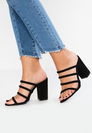 Personne portant un jean bleu et des sandales noires à talons hauts avec des lanières, debout sur une surface blanche.