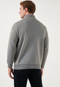 Felpa grigia con colletto alto e maniche lunghe, caratterizzata da una texture liscia e polsini a coste. Indossata con jeans scuri.