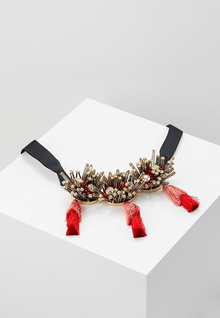 Collier décoratif avec des pointes dorées, des perles rouges, petits cristaux, pampilles rouges et une sangle noire réglable sur une surface blanche.