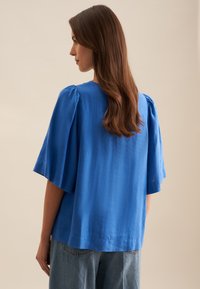 Top in lino blu con maniche corte a palloncino, vestibilità rilassata e collo rotondo. Abbinato a jeans in denim azzurro per un look casual.
