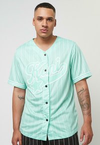 Minta zaļš pogājams krekls ar vertikālām baltām sv�ītrām, ar lielu baltu rakstītu logotipu un zvaigznītes detaļu priekšpusē. Īsas piedurknes.