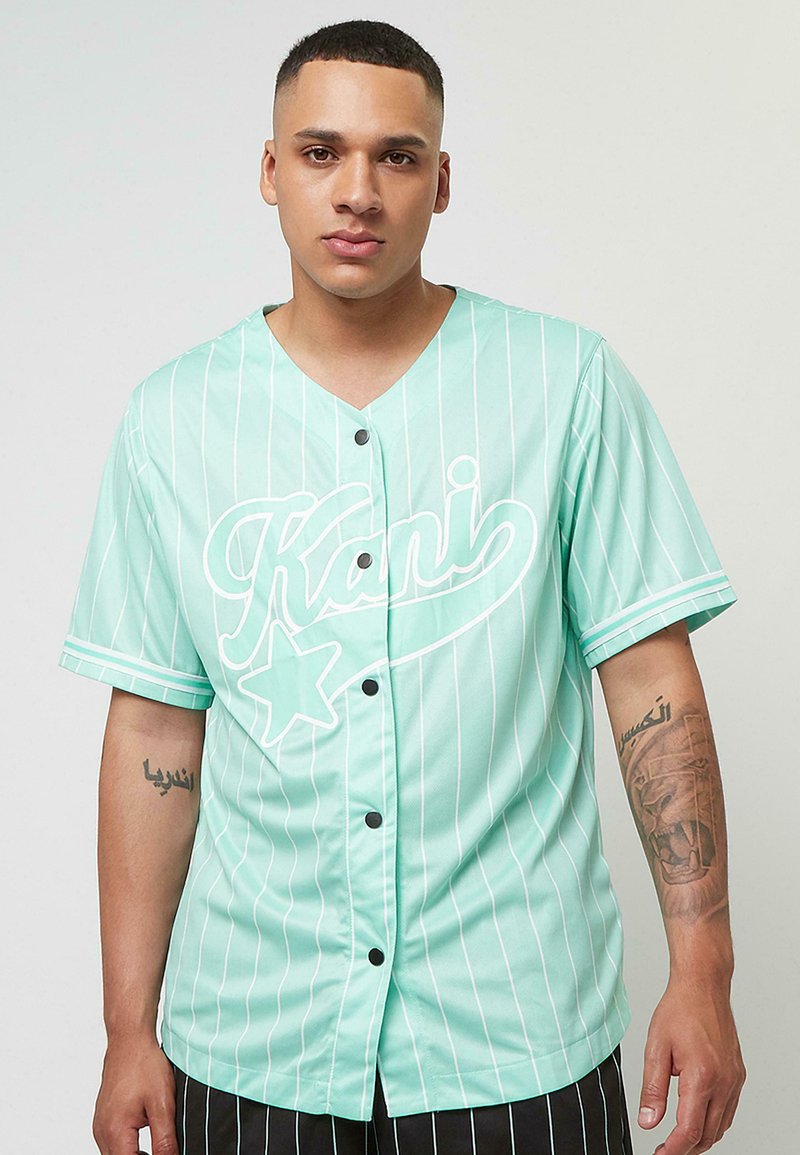 Minta zaļš pogājams krekls ar vertikālām baltām svītrām, ar lielu baltu rakstītu logotipu un zvaigznītes detaļu priekšpusē. Īsas piedurknes.