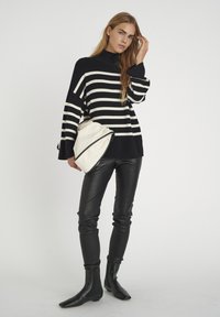 Gestreifter Pullover in Schwarz und Weiß mit hohem Kragen, kombiniert mit schwarzen Lederhosen und Stiefeletten, hält eine weiße Clutch.