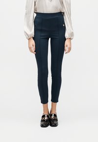 Γυναίκα που φοράει σκούρες μπλε παντελόνες μέχρι τον αστράγαλο με λεπτομέρειες ραφής μπροστά, ριγέ μπλούζα με μακριά μανίκια και μαύρα λουστρινένια μοκασίνια με χρυσές αλυσίδες.