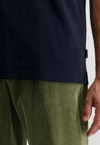 Close-up van een persoon die een marineblauwe shirt en groene corduroy broek draagt, met de rechterhand ontspannen langs de zijkant.