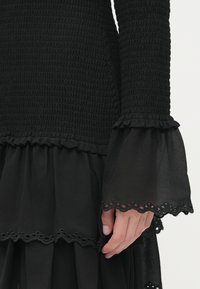 Mano descansando sobre un vestido negro con un corsé texturizado y mangas de campana transparentes con un borde de encaje en forma de concha.
