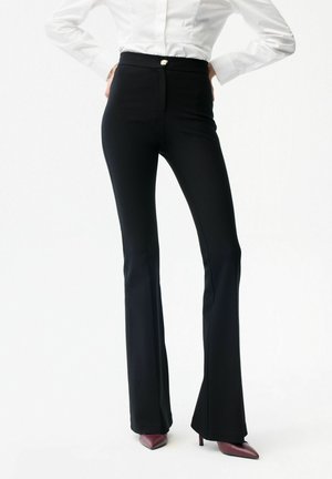 Femme portant un pantalon noir évasé taille haute avec un bouton doré, associé à des talons pointus bordeaux et un chemisier blanc.