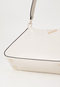 Sac à main synthétique blanc avec une forme structurée, quincaillerie dorée, garniture noire, et des clous décoratifs aux coins inférieurs.