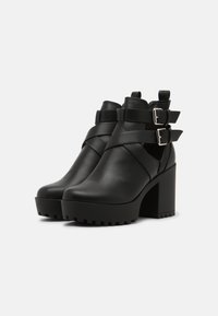 Botas negras de cuero hasta el tobillo con una plataforma gruesa y tacón alto en bloque. Presenta correas cruzadas y hebillas plateadas en el lateral. Textura suave.