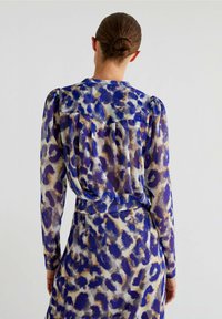 Blusa trasparente a maniche lunghe in stampa leopardata blu e beige, con spalle arricciate e dettaglio plissettato sul retro. Texture liscia con un design fluido.