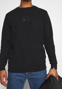 Sudadera negra de cuello redondo hecha de algodón con un logo tonal en el pecho, puños y dobladillo acanalados, combinada con jeans de mezclilla azul.