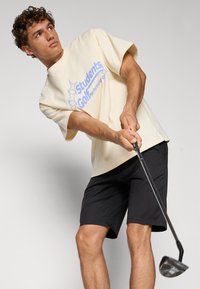 Beige bomull t-skjorte med blå grafisk tekst, kombinert med svarte shorts. Personen svinger en golfklubbe, og viser et avslappet atletisk utseende.