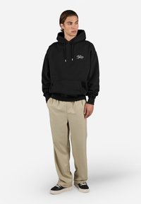 Black hoodie with white logo, kangaroo pocket, drawstrings, paired with beige trousers and black sneakers. Simple, relaxed fit.