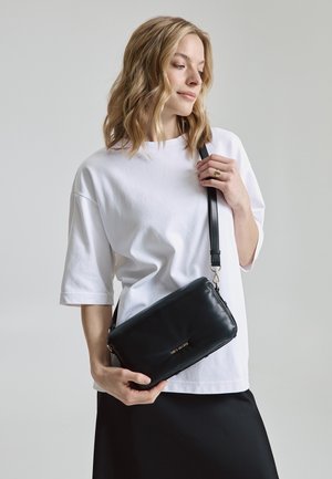 Schwarze Leder-Crossbody-Tasche mit schlanker Form, goldfarbenem Logo und verstellbarem Trageriemen, kombiniert mit einem weißen oversized T-Shirt und einem schwarzen Rock.