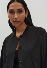 Schwarze Bomberjacke mit rundem Ausschnitt, Knopfverschluss und seitlichen Taschen. Glatte Textur in einfarbig und mit minimalen Details.