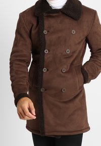 Brun dubbelknäppt kappa med fårskinnskrage, knapppdetaljer och slät tyg. Har långa ärmar och två sidofickor.