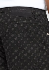 Jean en denim noir avec un motif subtil en gris foncé, accompagné d'une étiquette en cuir portant l'inscription "EIGHTYFIVE" sur la ceinture.