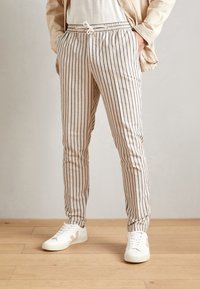 Pantaloni leggeri a righe in crema con strisce blu navy, dotati di vita elastica e cordoncino, abbinati a sneakers bianche su un pavimento in legno.