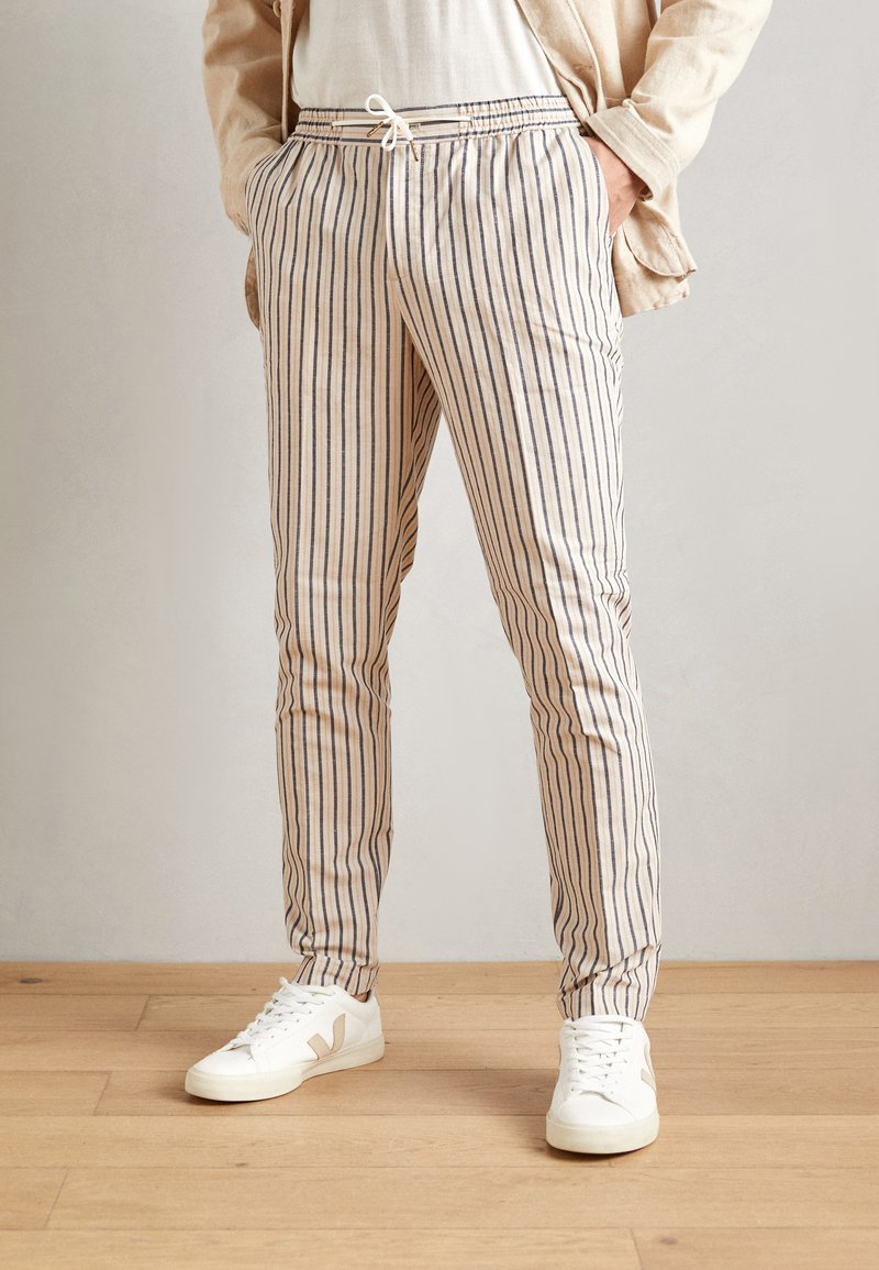 Pantaloni leggeri a righe in crema con strisce blu navy, dotati di vita elastica e cordoncino, abbinati a sneakers bianche su un pavimento in legno.