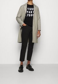 Light gray trench coat over a black t-shirt with graphic white text, paired with black trousers and black shoes with buckle accents.