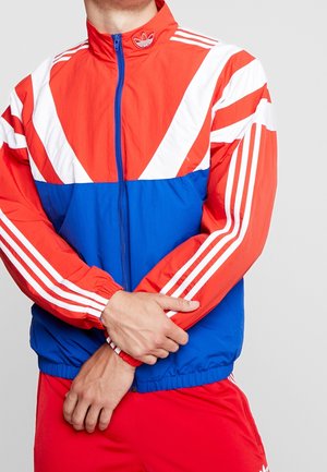 Mann trägt eine rot-weiß-blaue Adidas-Trainingsjacke mit Reißverschluss und weißen Streifen, hält einen Arm über seinen Körper.