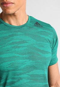 Zelené atletické tričko s kulatým výstřihem, texturovaným vlnovým vzorem, krátkými rukávy a černým logem Adidas na horní levé straně.