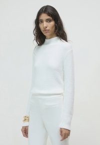 Maglione bianco strutturato con colletto alto e maniche lunghe. Abbinato a pantaloni bianchi aderenti. Un bracciale dorato al polso aggiunge un tocco metallico.
