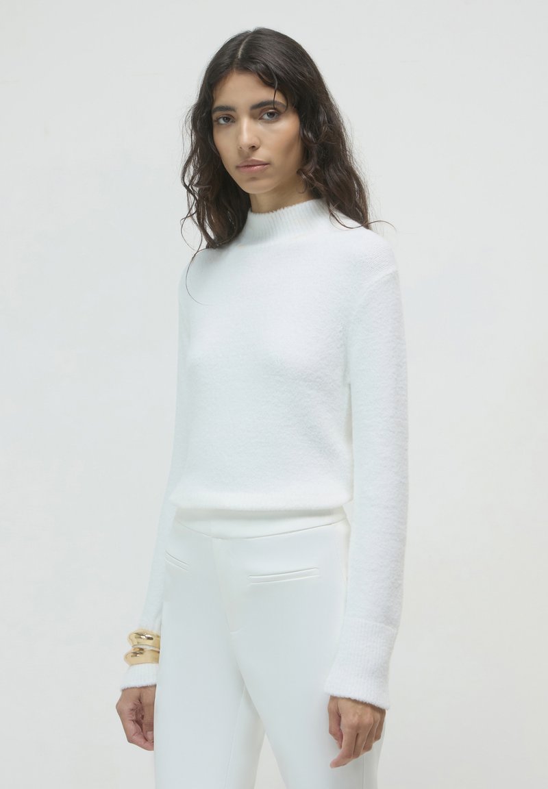 Maglione bianco strutturato con colletto alto e maniche lunghe. Abbinato a pantaloni bianchi aderenti. Un bracciale dorato al polso aggiunge un tocco metallico.