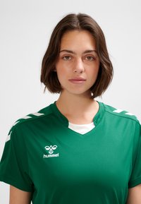 Green athletic shirt with a V-neck, made of smooth fabric. Features white Hummel logo and chevron accents on the shoulders.