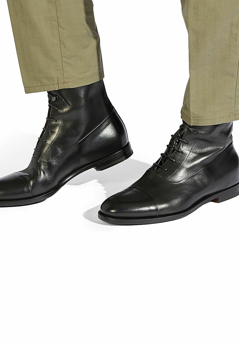 Botas de tobillo de cuero negro con un acabado pulido. Presentan un diseño de punta cap y cordones en la parte frontal, combinadas con pantalones caqui claros.