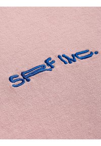 Różowy materiał o gładkiej fakturze z niebieskim haftowanym napisem "SRF Live". Centralny wzór, styl casualowy, bez dodatkowych wzorów czy akcentów.