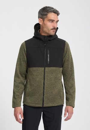 Mężczyzna w zielono-czarnej polarowej kurtce zapinanej na zamek z wysokim kołnierzem, patrzący na bok na jednolitym białym tle.