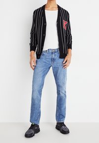 Cárdigan negro con rayas blancas y detalle en letras rojas, combinado con jeans de mezclilla azul claro y zapatillas deportivas negras.