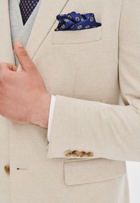 Beżowa marynarka o fakturze z wzorem w kratę, z kieszenią na piersi i niebieską poszetką w wzory. Guzik na mankiecie.