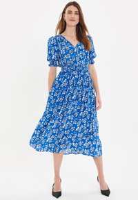 Vestido midi azul floral con mangas abullonadas, escote en V, cintura ajustada y falda fluida, con patrones de flores en blanco y negro. Tacones negros.