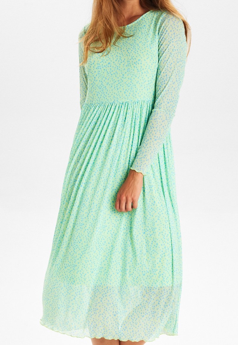 Robe verte menthe à manches longues avec un col rond, une jupe plissée et un petit motif floral bleu. Manches transparentes avec des poignets festonnés.