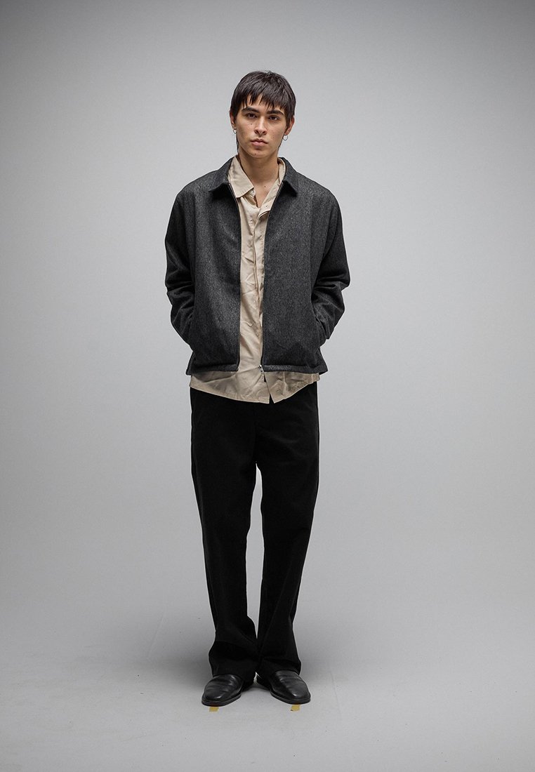 Νεαρός άντρας που στέκεται με τα χέρια στις τσέπες, φοράει σκούρο γκρι μπουφάν, μπεζ πουκάμισο, μαύρο παντελόνι και μαύρα παπούτσια, μπροστά σε απλό γκρι φόντο.