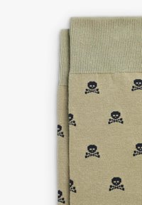 Calze al ginocchio di colore beige sabbia con una consistenza morbida, caratterizzate da un motivo ripetuto di teschi e tibie nere, e un bordo a costine verde in contrasto.