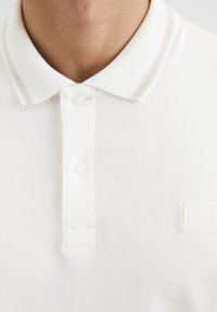 Weißes Poloshirt mit geripptem Kragen, der graue Streifen aufweist, drei Knöpfen und einem quadratischen Logopatch auf der Brust. Weiche Baumwolltextur.