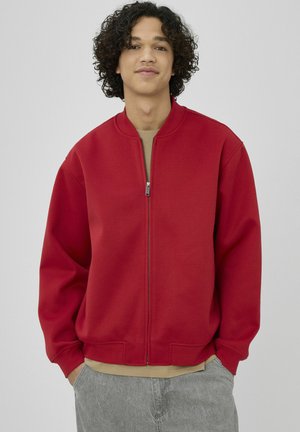 Veste zippée rouge avec col arrondi, poignets côtelés et coupe décontractée, superposée à une chemise beige, sur un fond uni.