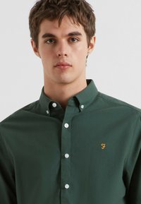 Donkergroene katoenen overhemdbloes met een kraag en witte knopen. Inclusief een klein geel logo aan de linkerkant.