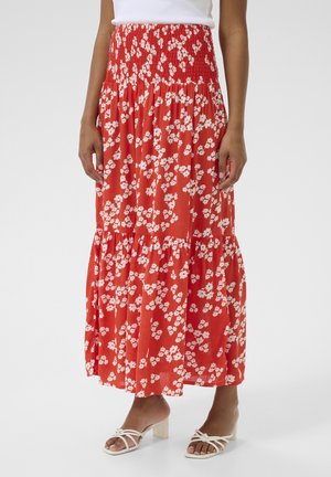 Maxi skirt in red fabric with white floral pattern, featuring a smocked waistband and tiered design. Worn with white strappy sandals.