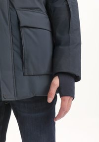 Jūrų laivyno išorinis švarkas su paminkštinta konstrukcija, turintis priekinę kišenę, reguliuojamus rankogalius ir tekstūruotą megztą sluoksnį ties riešu.